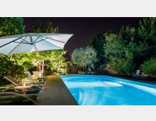 Piscina all’aperto illuminata di notte, con acqua azzurra e bordo in cemento. Lungo il lato ci sono lettini prendisole sotto un grande ombrellone bianco, circondati da alberi e vegetazione fitta.