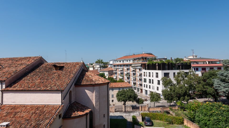 Vista urbana con tetti in coppi di un edificio storico in primo piano e palazzi residenziali moderni sullo sfondo. In basso si vedono alberi, un’area verde e una strada con parcheggi sotto un cielo sereno.