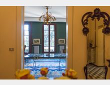 Vista attraverso una porta su un salotto interno con pareti verde scuro, grandi finestre a vetri con cornici in legno e un lampadario centrale. Al centro c’è un tavolo con piano in vetro su un tappeto a motivi geometrici, con termosifoni e quadri alle pareti; a destra si vede un corridoio con pavimento a scacchi e un attaccapanni in metallo decorato.