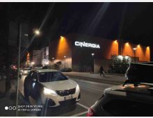 Veduta notturna dell’esterno di un moderno cinema con grande insegna luminosa “CINERGIA” su una facciata arancione illuminata. In primo piano si vede una strada con auto parcheggiate e lampioni lungo il marciapiede.