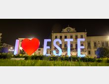 Inquadratura notturna di una piazza con una grande installazione luminosa “I ♥ ESTE” davanti a un’aiuola fiorita. Sullo sfondo si vede un edificio storico con facciata simmetrica, balconi e un orologio centrale in sommità, illuminato da lampioni.