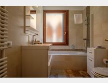 Bagno moderno con piastrelle beige, lavabo con mobile sospeso e specchio, vasca da bagno con doccetta e fascia a mosaico. Una finestra con vetro satinato e cornice in legno illumina l’ambiente; a lato si vedono scaldasalviette e mobiletto con cassetti.