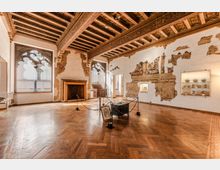 Sala museale in un palazzo storico con pavimento in parquet a spina di pesce e soffitto a travi lignee decorate. Alle pareti, intonaco parzialmente scrostato con affreschi visibili, grandi finestre ad arco, un camino in pietra e vetrine illuminate con ceramiche esposte.