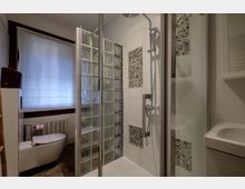 Bagno moderno con box doccia in vetro e parete divisoria in mattoni di vetro, rivestimenti chiari con inserti a mosaico di ciottoli. A sinistra si vede un WC sospeso sotto una finestra con tenda, a destra un lavabo con mobile.