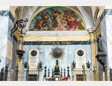 Interno di una cappella monumentale con pareti in marmo policromo, cancellata in ferro battuto e altare centrale con candelabri e decorazioni. Sopra l’altare si vede un affresco semicircolare con figure angeliche e una scritta dorata, affiancato da lapidi commemorative e statue laterali.