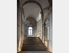 Interno di un edificio storico con ampia scalinata in pietra che sale verso una finestra ad arco. Volte a botte, colonne e cornici in pietra incorniciano pareti in muratura a vista e intonaco, con corrimano metallici e illuminazione discreta lungo i lati.