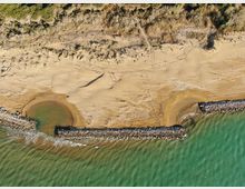 Veduta dall’alto di una spiaggia sabbiosa con dune e vegetazione retrostante, con rami e legni sparsi sulla sabbia. In primo piano una scogliera frangiflutti in pietra forma due piccole insenature davanti al mare verde.