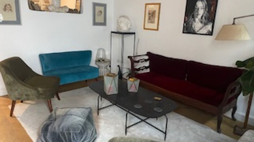 Soggiorno con pareti bianche arredato con divano bordeaux, divanetto blu e poltrone, centrato su un tavolino basso in metallo su tappeto chiaro. Alle pareti sono appesi quadri e una lampada da terra illumina l’angolo vicino a una pianta.