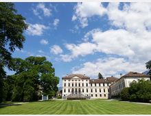 Ampio prato con alberi che conduce a una grande villa storica color crema, con facciata simmetrica, finestre ornate e scalinata centrale. Sullo sfondo cielo azzurro con nuvole e ali laterali dell’edificio con tetti in tegole rosse.