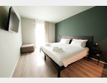 Camera da letto moderna con letto matrimoniale in lenzuola bianche e testiera scura, parete verde e pavimento in legno chiaro. Di fronte al letto ci sono una TV a parete e una panca imbottita, con una grande finestra a tutta altezza con tende che illumina l’ambiente.