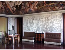Interno di una sala storica con pavimento scuro, divanetti e tavolo in legno, e una grande parete decorata con un bassorilievo monocromo in stile classico. Il soffitto è affrescato con scene figurative e una cornice ornamentale corre lungo il bordo superiore delle pareti.