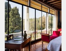 Interno di una camera con pavimento in legno e soffitto con travi a vista, affacciata su una lunga parete di finestre a tutta altezza con telai scuri che danno su una terrazza con balaustra e colonne. Nell’ambiente si vedono una scrivania in legno, una poltrona rossa e un letto, con vista su alberi e colline all’esterno.