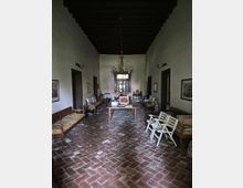 Ampio salone interno con pavimento in mattoni a spina di pesce, pareti intonacate chiare e soffitto alto con travi in legno a vista. Al centro ci sono un tavolo e un lampadario, con panche e sedie lungo i lati e una porta vetrata in fondo alla stanza.