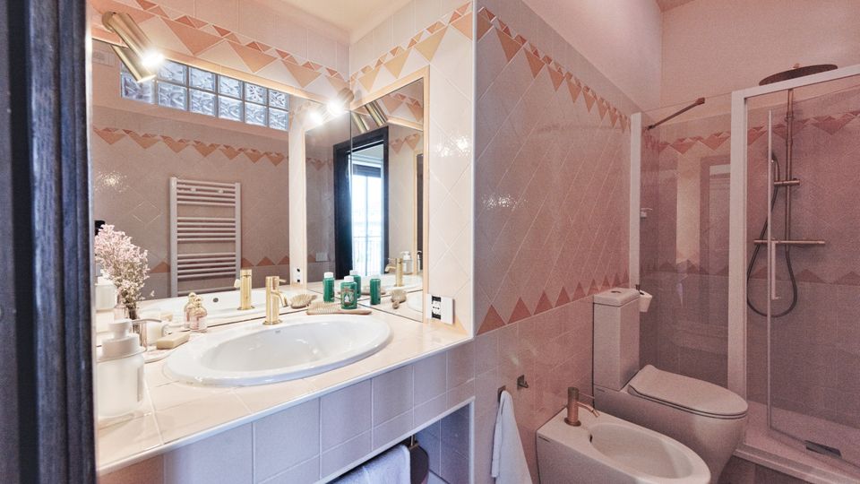 Bagno moderno con rivestimento in piastrelle chiare e decoro geometrico, lavabo incassato su ampio piano con grande specchio e luci. A destra si vedono bidet e WC e una doccia angolare con box in vetro.