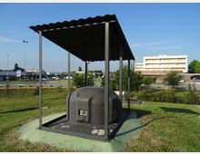 Struttura metallica con tettoia a falda singola che copre una cupola in cemento con una piccola apertura, situata su un prato lungo una strada. Sullo sfondo si vedono parcheggi, lampioni e un edificio commerciale a più piani.