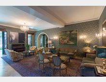 Salotto elegante in stile classico con carta da parati damascata, grandi tappeti persiani e numerose sedute attorno a tavolini in vetro. Lampadari e applique illuminano la stanza, con un pianoforte, quadri alle pareti e un arco che conduce ad altre stanze.