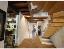 Interno di un’abitazione con pareti bianche, soffitto con travi in legno a vista e pavimento in parquet a spina di pesce. A destra una scala moderna sospesa in legno e, a sinistra, una libreria a tutta altezza con libri e oggetti decorativi.