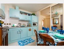 Cucina abitabile con mobili azzurro chiaro, piano di lavoro con fornelli e elettrodomestici integrati, e tavolo da pranzo in legno apparecchiato con piatti turchesi e bicchieri. Sul lato destro è presente una vetrata decorativa che separa l’ambiente da un corridoio interno.