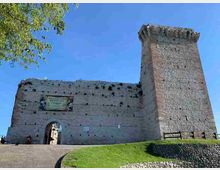 Fortezza in pietra su un terrapieno erboso, con mura alte, ingresso ad arco e una torre quadrata merlata sulla destra. In primo piano c’è un vialetto in salita e sullo sfondo un cielo azzurro con rami di albero in alto a sinistra.