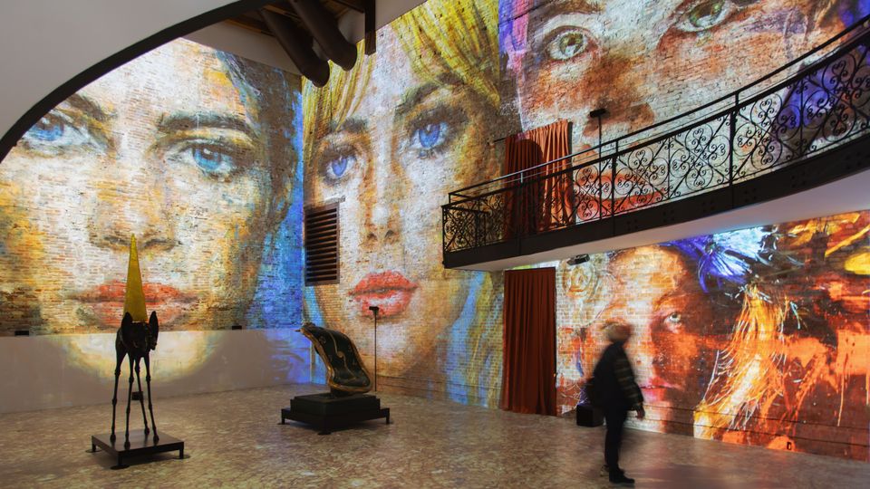 Interno di una galleria o museo con ampie pareti in mattoni su cui sono proiettati grandi ritratti colorati che coprono quasi tutta la superficie. Lo spazio ha un soppalco con ringhiera in ferro battuto, tende rosse e alcune sculture esposte su piedistalli sul pavimento in pietra.