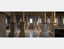 Ampia sala interna in pietra con file di colonne e pilastri massicci su basamenti quadrati, soffitto a travi e pavimento in lastre. Sul fondo si vedono grandi finestre ad arco che lasciano entrare luce naturale tra le arcate.