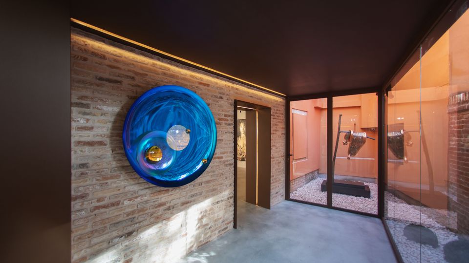 Interno di uno spazio espositivo moderno con parete in mattoni a vista e grande installazione circolare in vetro blu, pavimento in cemento e soffitto scuro. Sulla destra, una parete vetrata affaccia su un piccolo cortile con ghiaia e un’opera scultorea metallica su base nera.