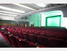 Interno di una sala cinematografica con file di poltrone rosse numerate disposte in pendenza verso un grande schermo incorniciato e illuminato. Soffitto curvo con luci a neon, pareti con rivestimento in legno e balconate laterali.