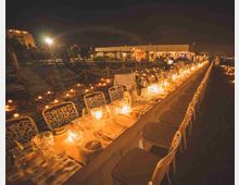 Lunga tavolata all’aperto su una spiaggia di notte, apparecchiata con tovaglie chiare, sedie in ferro decorato e numerose candele che illuminano il percorso. Sullo sfondo si vedono strutture e un edificio illuminati, con lanterne e luci sparse sulla sabbia.