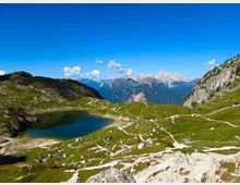 Paesaggio montano con un piccolo lago alpino incastonato tra prati verdi e rocce chiare, attraversati da sentieri sterrati. Sullo sfondo si vedono cime rocciose e una parete scoscesa sotto un cielo limpido.