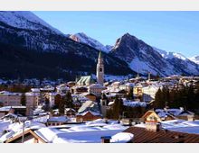 Panorama di un paese alpino innevato con tetti coperti di neve e una grande chiesa con alto campanile al centro. Sullo sfondo si vedono montagne ripide e boscose con cime innevate sotto un cielo sereno.
