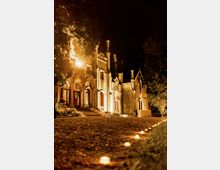 Facciata di un grande edificio storico illuminato di notte, con archi d’ingresso, finestre alte e numerosi comignoli sul tetto. Un vialetto in ghiaia con piccole luci a terra conduce ai gradini, fiancheggiato da alberi e vegetazione.