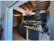 Cucina moderna open space con isola centrale e bancone in legno, mobili scuri e piano di lavoro attrezzato. Soffitto con travi a vista e luci a sospensione, con passaggio ad arco verso un’altra zona della casa.