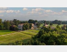 Veduta aerea di una villa storica con edifici in pietra e tetti in coppi, circondata da ampi prati e alberi. Un lungo viale attraversa il parco e conduce al complesso, con campi e boschi sullo sfondo sotto un cielo parzialmente nuvoloso.