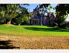Edificio storico in stile neogotico, simile a un castello, con archi e torrette, visto in fondo a un ampio prato verde. In primo piano si nota un tappeto di foglie autunnali e grandi alberi che incorniciano la scena.