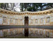 Esedra semicircolare in pietra decorata con bassorilievi e nicchie con figure scolpite, con un’arcata centrale e gradini alla base. Davanti si trova una vasca d’acqua che riflette la facciata, circondata da alberi sullo sfondo.