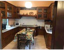 Cucina tradizionale con mobili in legno scuro e pensili con ante in vetro, disposta a U con ampi piani di lavoro e lavabo a destra. Al centro c’è un tavolo da pranzo con tovaglia a fantasia, sotto un lampadario a globo, con pareti piastrellate chiare e una finestra sul lato sinistro.