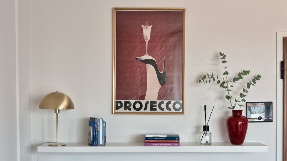 Parete interna di un ambiente domestico con mensola bianca sopra un radiatore, decorata con lampada da tavolo in ottone, libri, diffusore e vaso rosso con rami verdi. Sopra la mensola è appeso un grande poster incorniciato con scritta “PROSECCO” su fondo rosso.