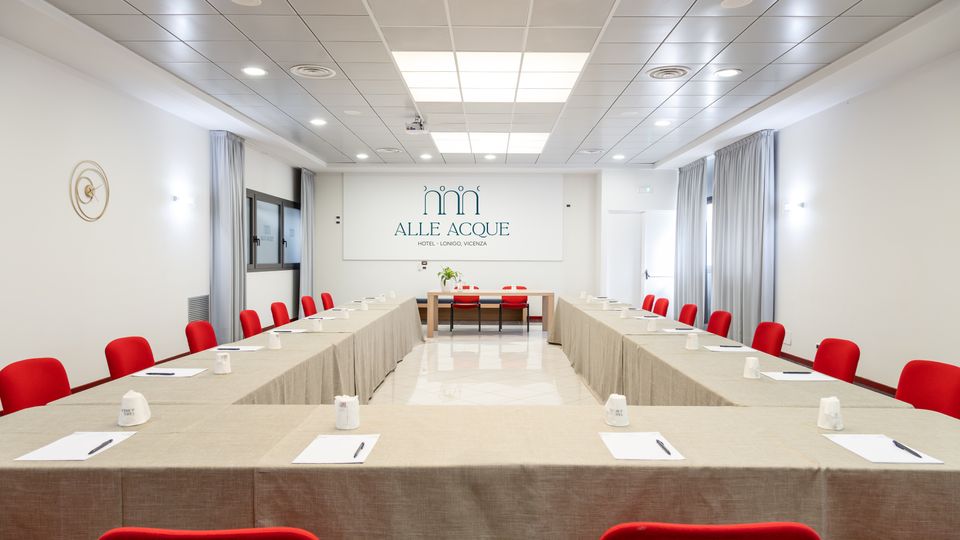 Sala conferenze moderna con tavoli disposti a U coperti da tovaglie beige, sedie rosse e blocchi per appunti. Soffitto a pannelli con illuminazione incassata, pareti bianche con tende grigie e parete frontale con insegna “Alle Acque”.