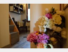 Interno di una sala da pranzo domestica con tavolo apparecchiato e sedie, pavimento in legno e scala laterale; sullo sfondo una mensola con oggetti. In primo piano, un grande bouquet di fiori artificiali bianchi e rosa appoggiato contro una parete.