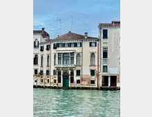 Facciata di edifici storici affacciati su un canale, con intonaco chiaro parzialmente scrostato, tetti in coppi rossi e persiane scure. Al centro si vede un portale ad arco con balcone e finestre ad arco, mentre in primo piano scorre l’acqua del canale.