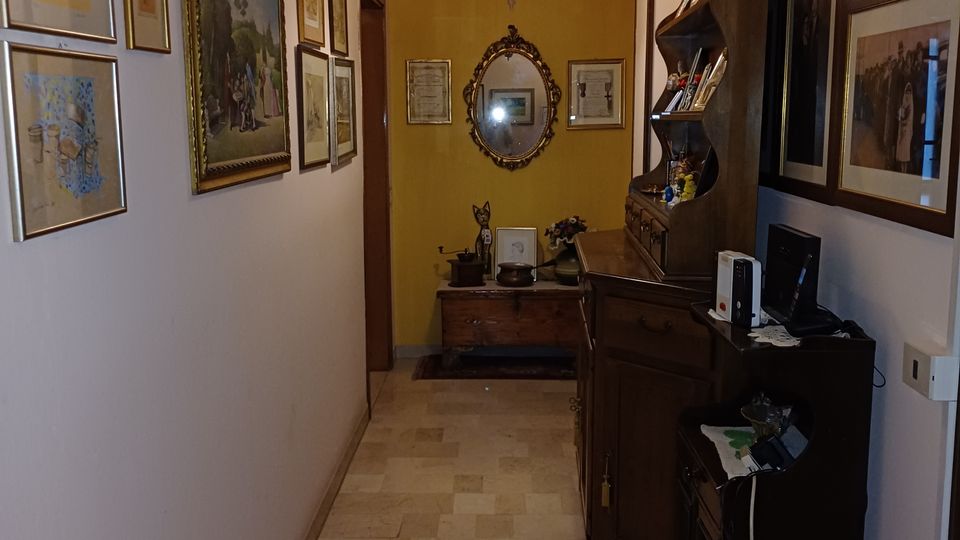 Corridoio interno di una casa con pavimento in pietra chiara, pareti decorate da numerosi quadri incorniciati e lampadario a sospensione. Sul lato destro ci sono mobili in legno con oggetti e una specchiera ovale sulla parete di fondo.
