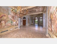 Ampia sala storica con pavimento in seminato veneziano, pareti affrescate con scene figurative e colonne effetto marmo. Soffitto decorato con riquadri dipinti, grandi lampadari in ferro battuto e alte finestre ad arco che si aprono su un balcone con vista sul verde.