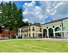 Complesso di edifici in stile villa con facciate colorate e tetti in coppi, affacciato su un prato verde con un grande albero. A destra si vede una struttura con ampie arcate e finestre circolari, con area esterna arredata con tavoli e ombrelloni.