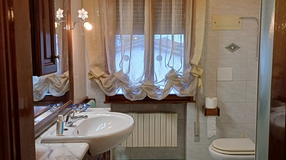 Bagno stretto con pavimento verde e pareti piastrellate chiare, mobile lavabo in legno con specchiera illuminata. In fondo una finestra con tende leggere e termosifone sotto, e sul lato destro WC e bidet accanto a una doccia con parete in vetro.