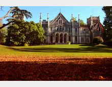 Facciata di una villa storica in stile neogotico, con archi, finestre alte e torrette, affacciata su un ampio prato. L’edificio è circondato da grandi alberi e vegetazione, con cielo sereno sullo sfondo.