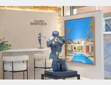 Interno di una galleria d’arte con pareti bianche e insegna “Galeries Bartoux”, bancone con sgabelli e una scultura in primo piano su piedistallo. Sullo sfondo è appeso un grande quadro raffigurante una villa moderna con piscina e palme.