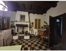 Interno rustico con soffitto a travi in legno e pavimento a scacchi bianco e nero, dominato da un grande camino in muratura con mensola decorata. Sulla destra ci sono una finestra con tende e un tavolo da lavoro inclinato con oggetti, mentre una porta scura aperta conduce a un’altra stanza.