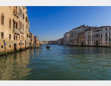 Ampio canale cittadino fiancheggiato da palazzi storici con facciate chiare, finestre ad arco e balconi, con pali d’ormeggio lungo le rive. Acqua calma al centro del canale sotto cielo sereno, con una gondola in lontananza.
