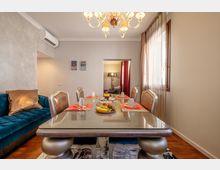 Sala da pranzo in un appartamento, con grande tavolo in vetro apparecchiato e sedie imbottite, lampadario a soffitto e divano in velluto blu sul lato sinistro. Ampia finestra con tende leggere a destra e porta sul fondo che conduce a un’altra stanza.