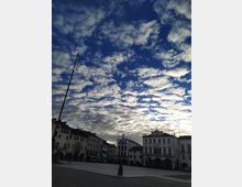 Ampia piazza cittadina con edifici storici a più piani e portici, tra cui un palazzo con orologio sulla facciata. In primo piano una pavimentazione in pietra con un lampione e un alto pennone, sotto un cielo molto esteso ricco di nubi.